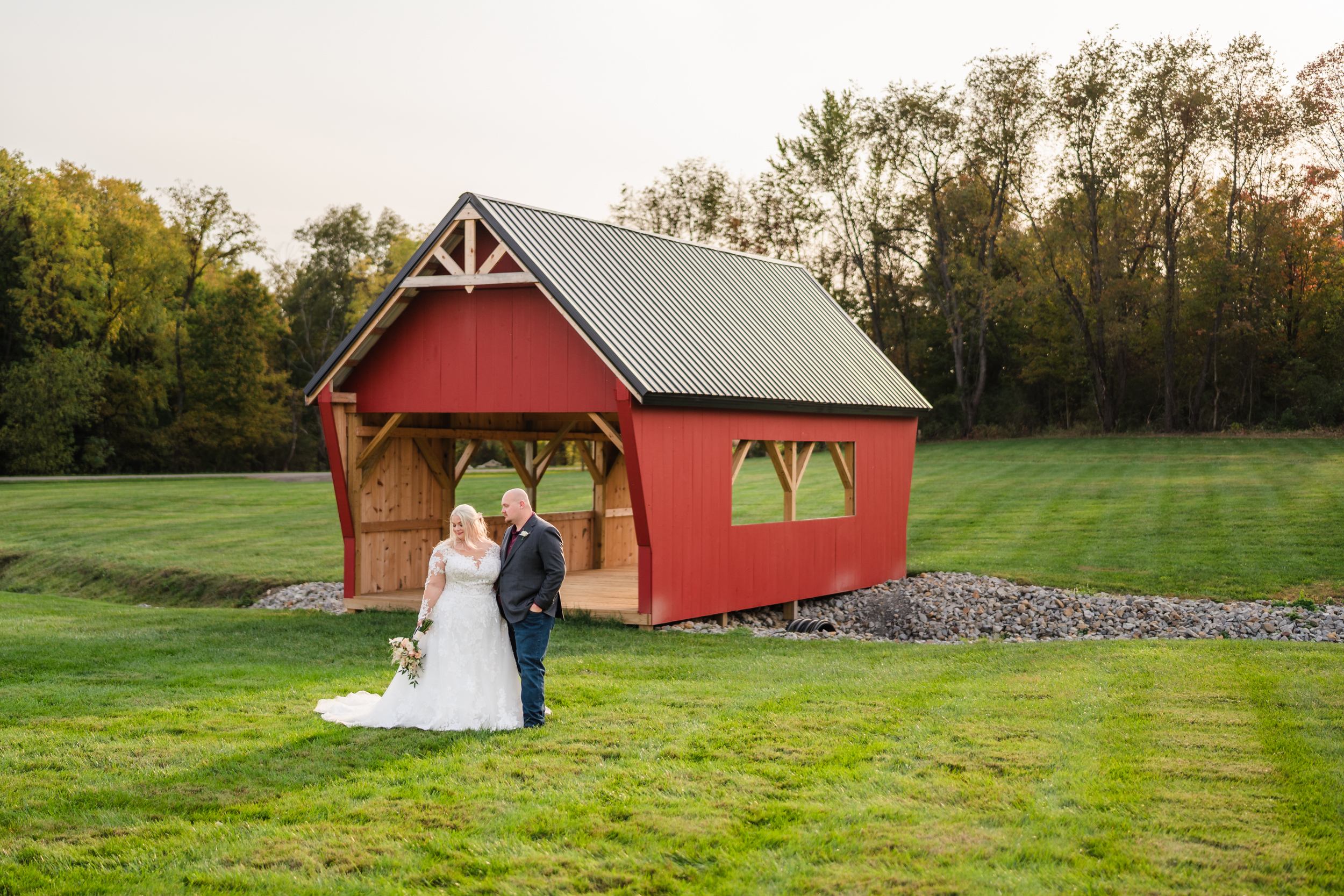 A groom smiles at his bride as they stand in front of a red covered bridge on the lawn of their rustic wedding venue