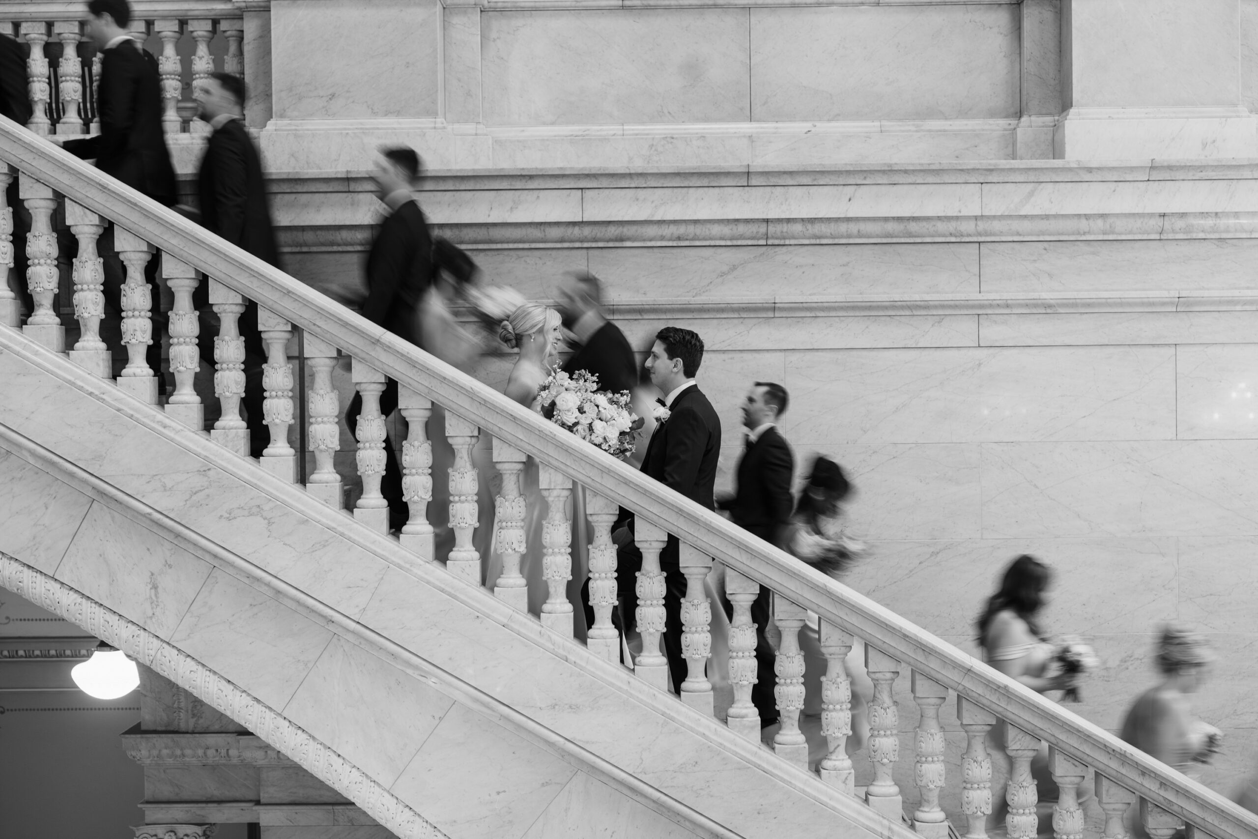 A bride and groom smile at each other on marble steps inside the Ohio Statehouse while their wedding party walks behind them